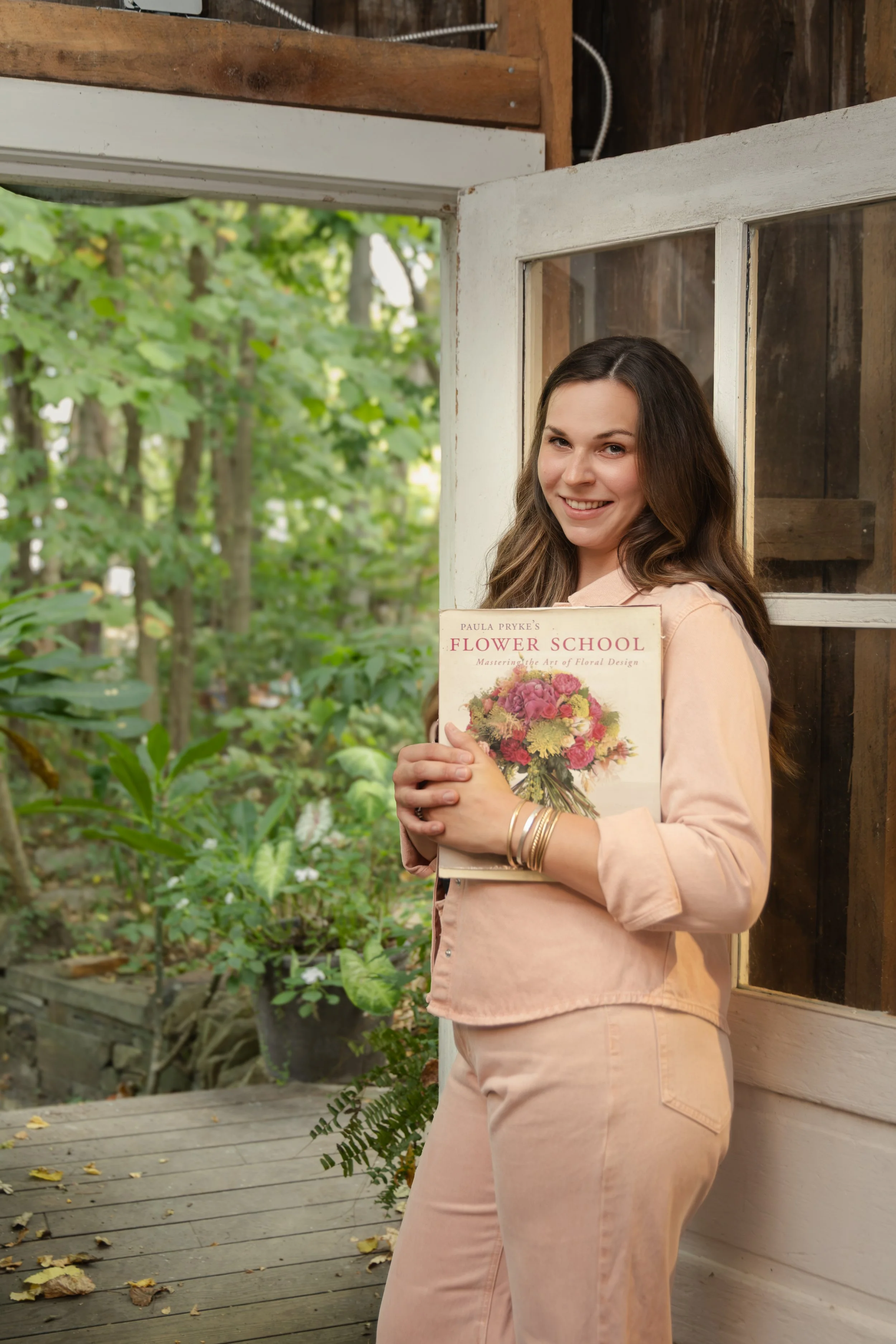 A woman standing outside on a wooden porch, holding a book titled 'Flower School' with a floral cover, smiling, with greenery in the background.