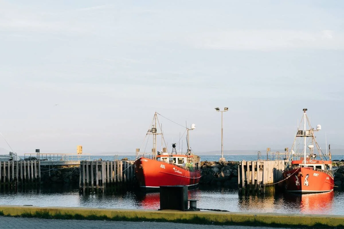 Two red boats docked at a marina, with a breakwater and cloudy sky in the background.