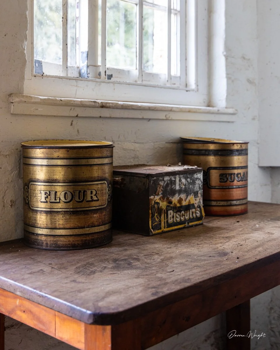 Three vintage metal containers labeled 'FLOUR,' 'Biscuits,' and 'SUGAR' sit on a wooden table near a window in a rustic kitchen.