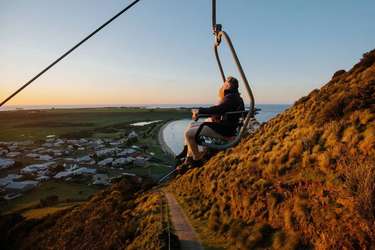 A person sitting on a scenic sky lift chair overlooking a coastal town at sunset, with hills and water in the background.