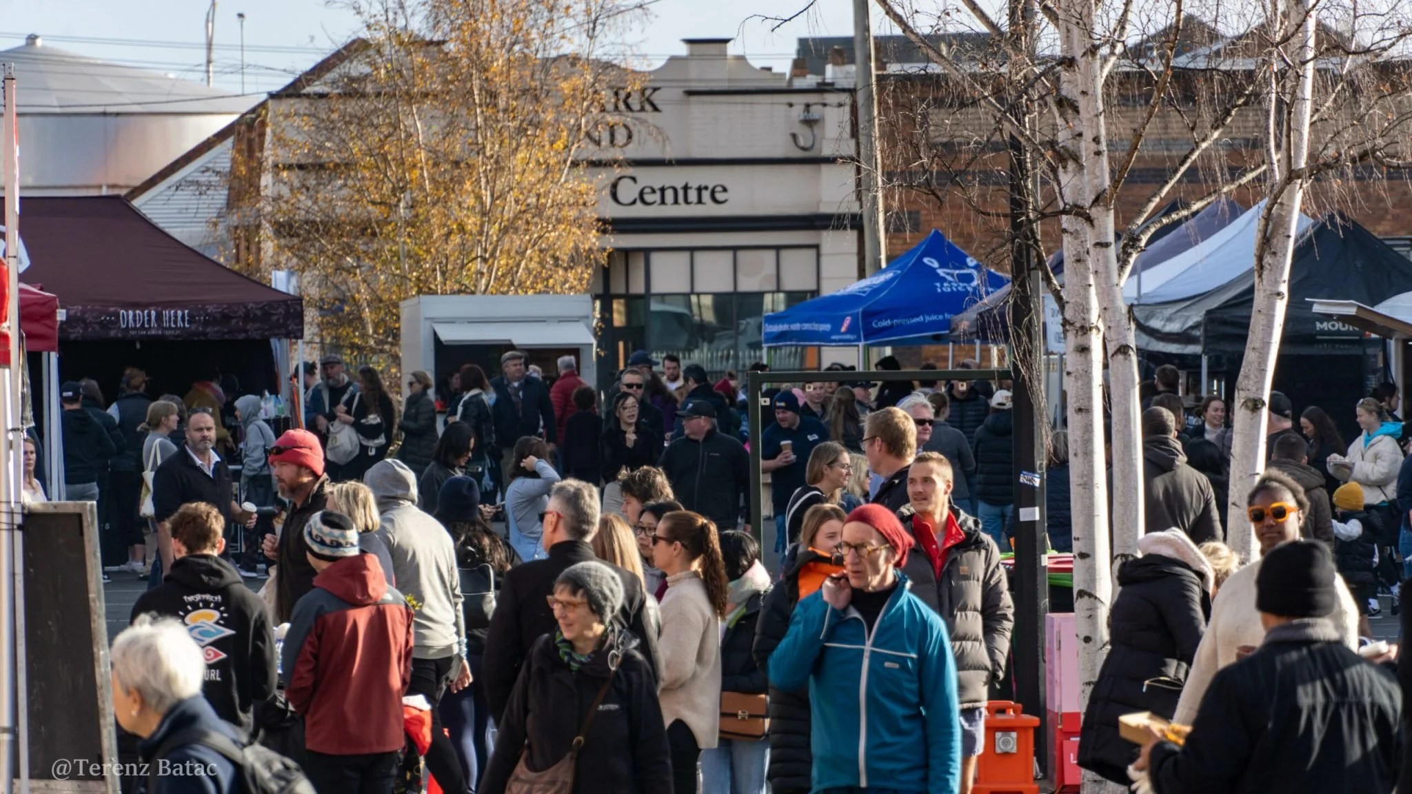 Crowd of people at an outdoor market with vendor tents and trees.