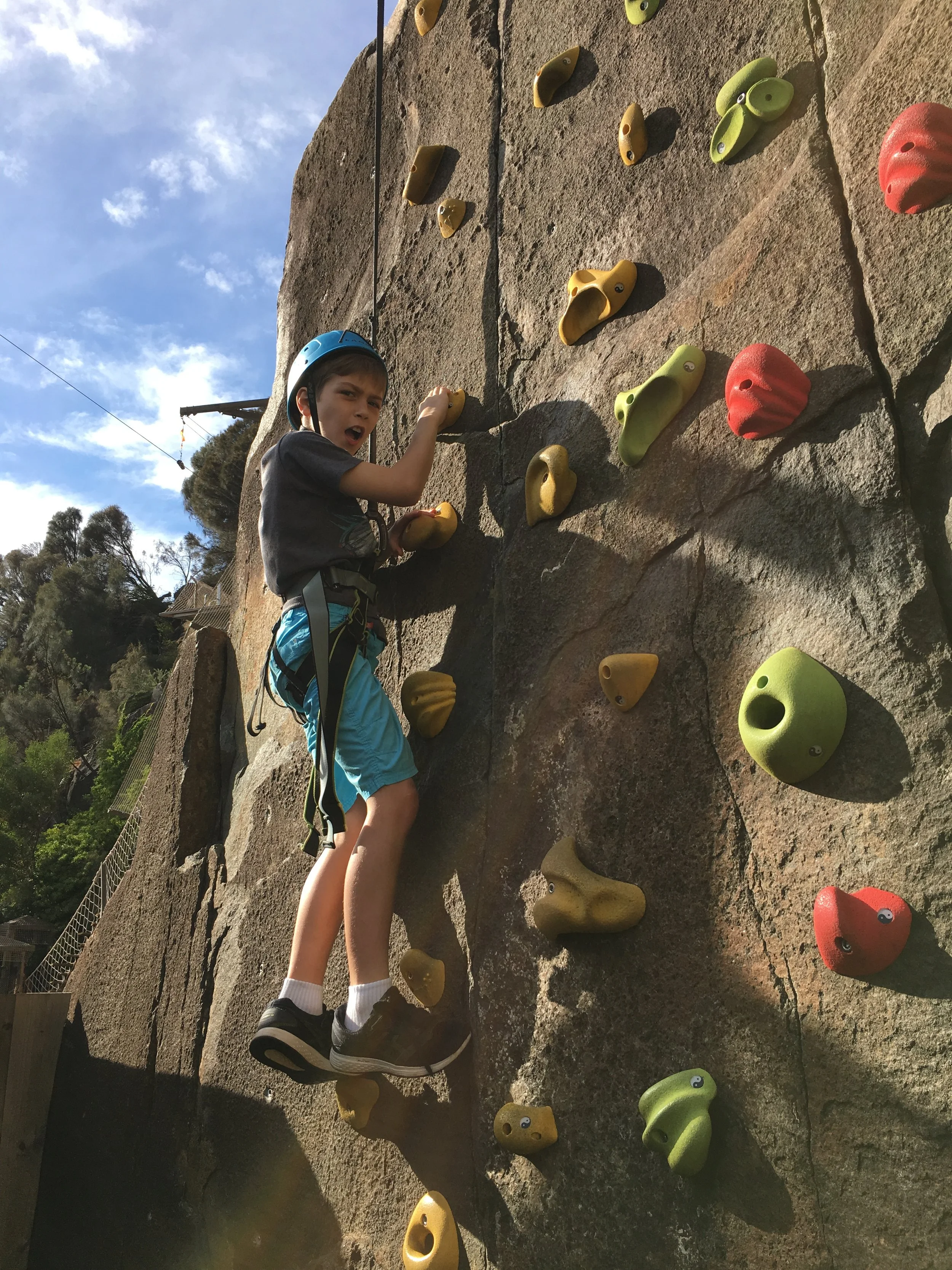 A boy wearing a blue helmet, gray shirt, and blue shorts climbing a rock wall outdoors with colorful handholds and footholds.