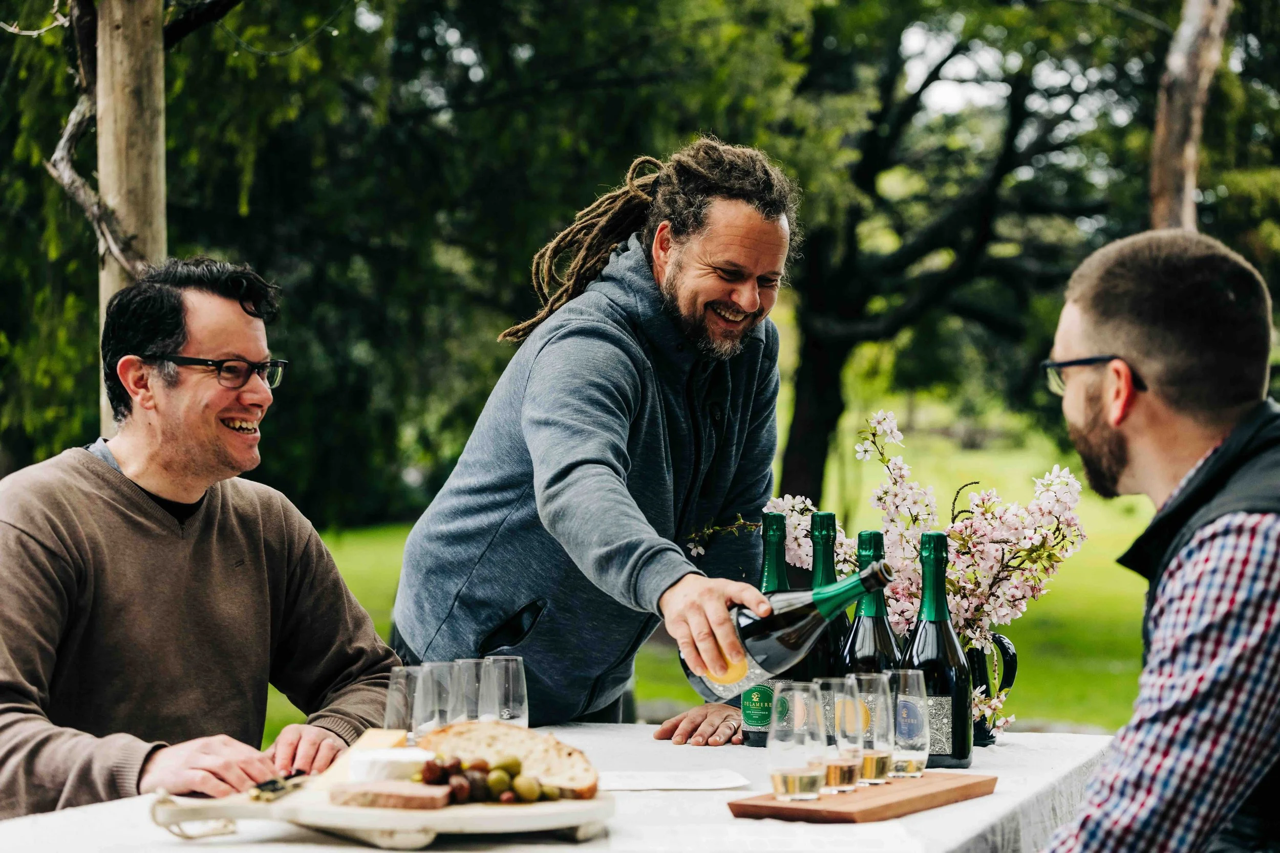 Three men having a outdoor celebration, with one pouring sparkling wine into glasses on a table with food and flowers.