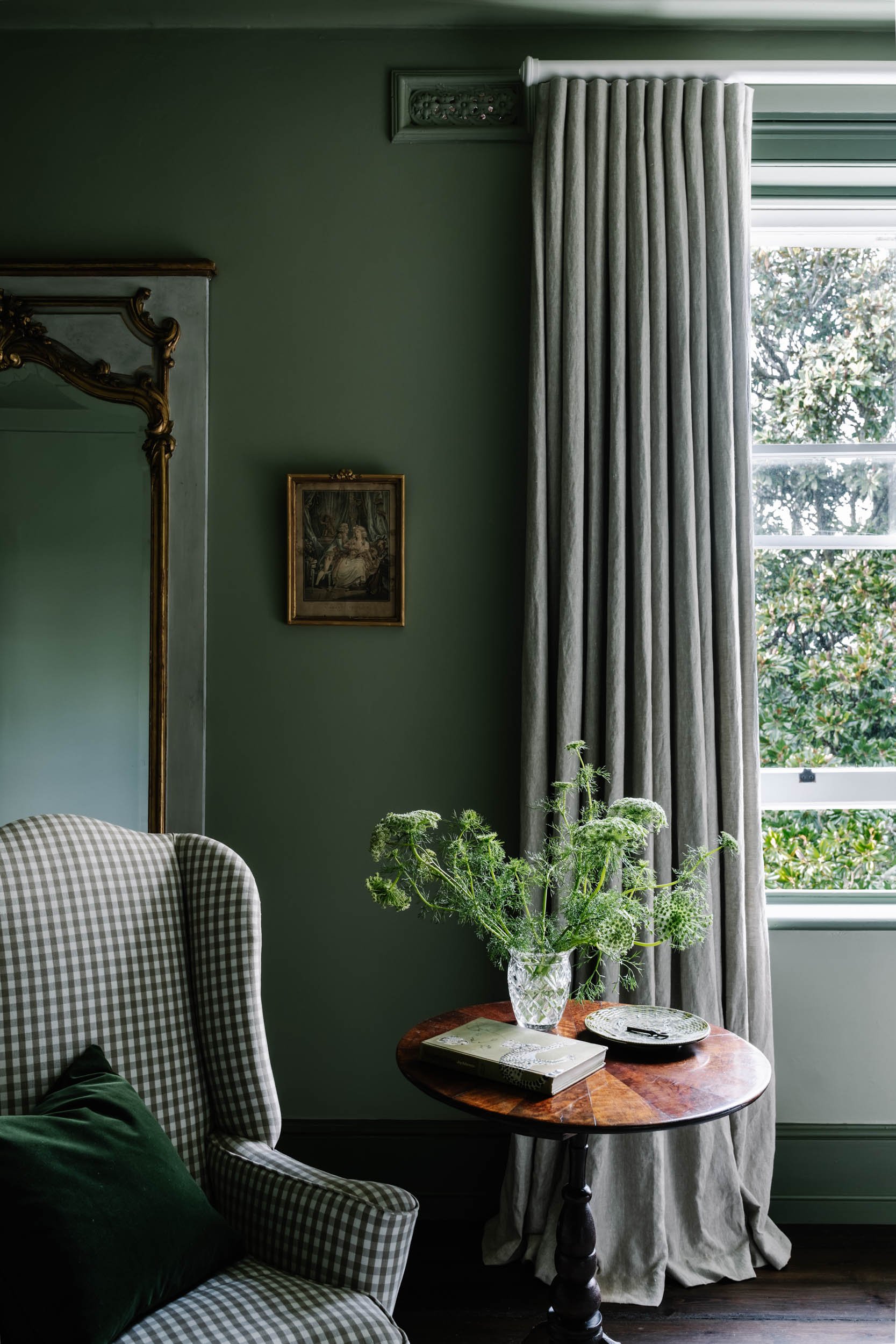 A cozy corner of a room with a checkered armchair, a small wooden side table with a glass vase of greenery, a book, and a plate, a window with striped curtains, green walls, and a small painting on the wall.