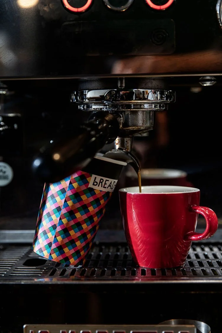 Coffee brewing from an espresso machine into a red mug, with a colorful patterned cup labeled 'BREAD' in place of a filter.
