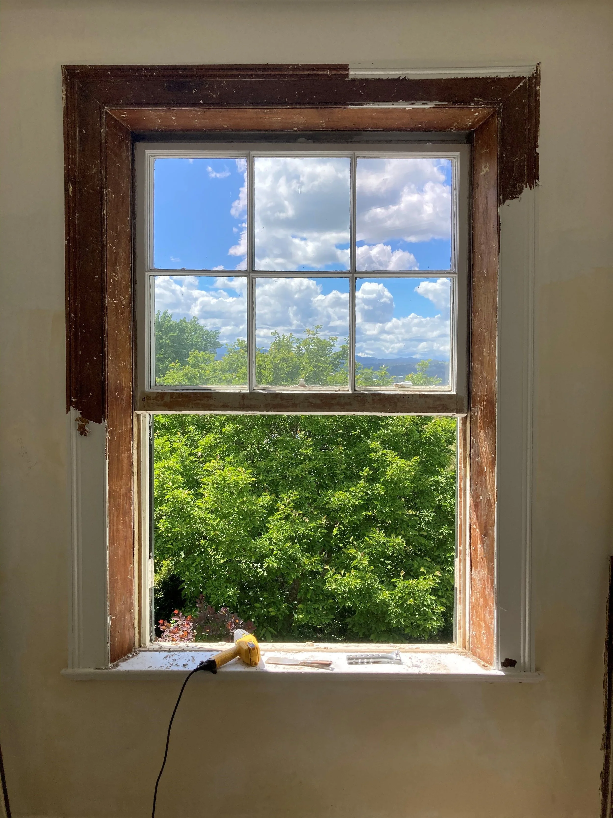 View through a window showing a bright blue sky with fluffy white clouds and green trees outside, with tools and a yellow drill resting on the windowsill inside the room.