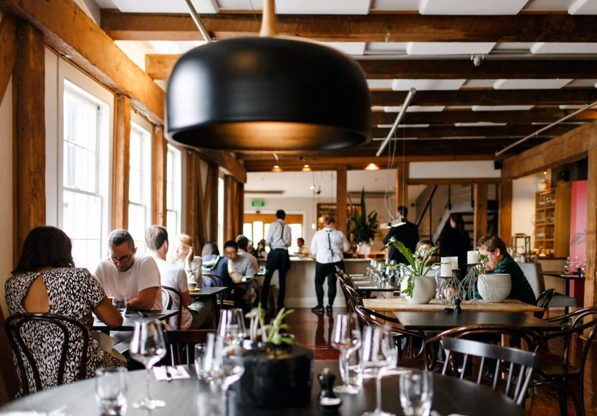 Interior of a restaurant with wooden beams, large windows, and several tables occupied by customers dining. A large black pendant light hangs from the ceiling, and a bar area is visible in the background. The overall ambiance is warm and inviting.
