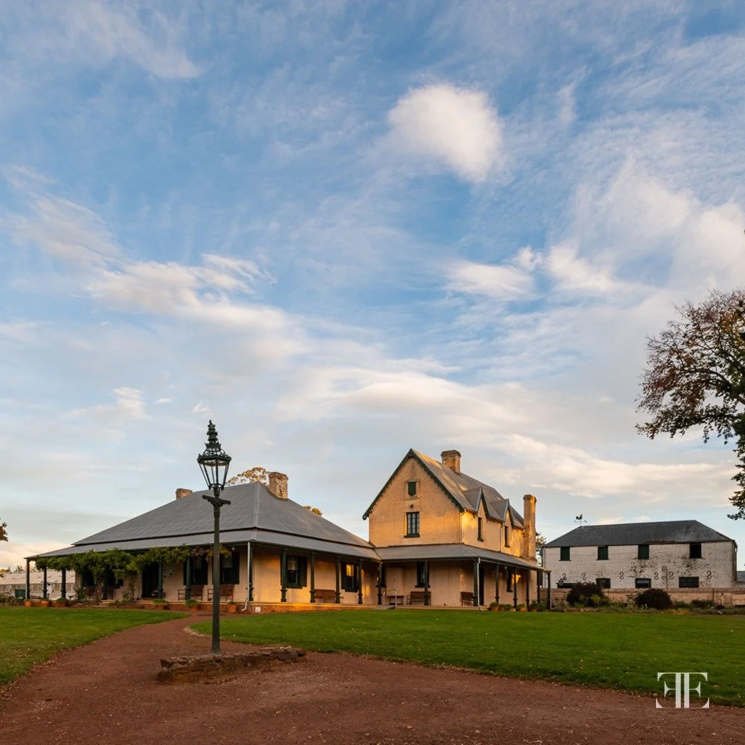 Historic two-story house with a veranda, surrounded by a grassy yard and a dirt path, under a partly cloudy sky with a vintage streetlamp nearby.