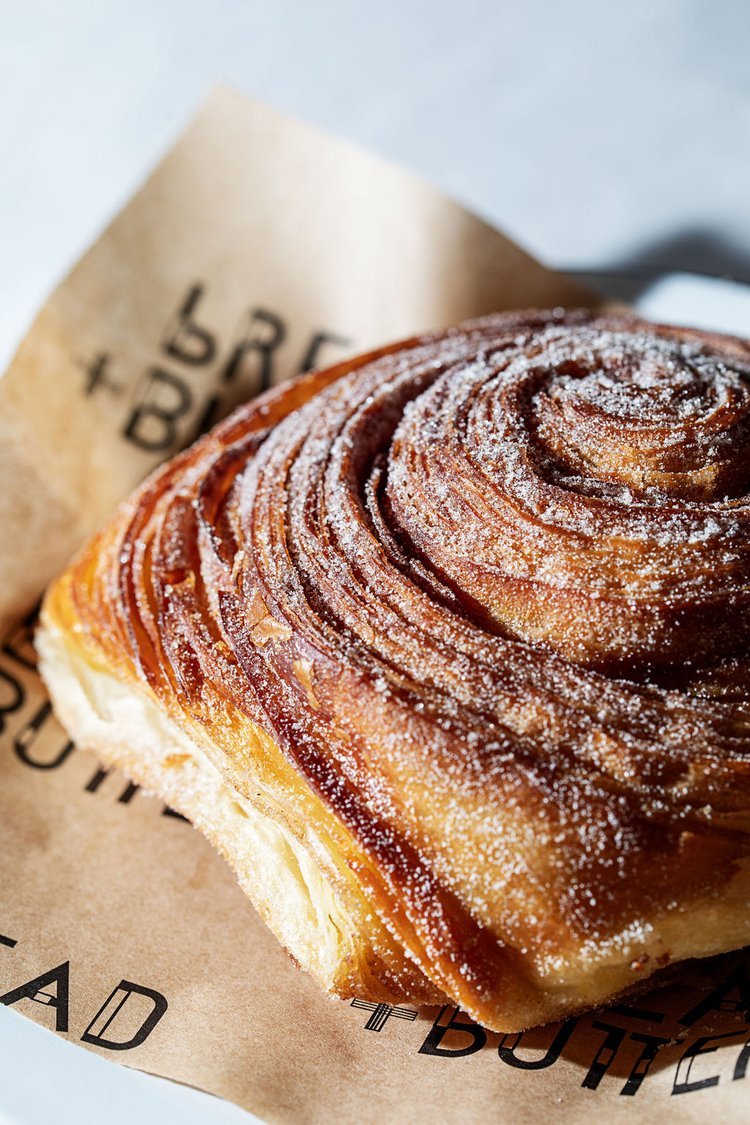 Close-up of a cinnamon roll dusted with powdered sugar on parchment paper that has some text, with a blurred background.