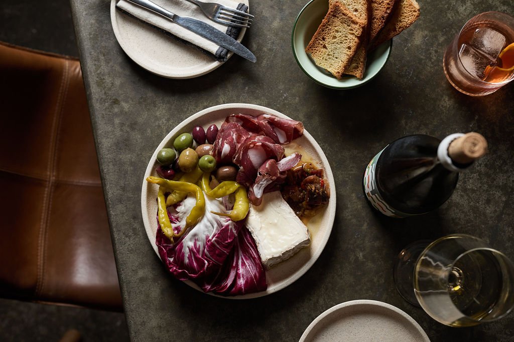 A plate of assorted food including grapes, prosciutto, cheese, and vegetables on a dark table with bread, a glass of wine, and a bottle of wine nearby.