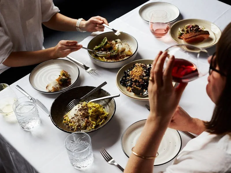 People enjoying a meal; one person drinking red wine, another using a fork and knife at a table with various plates of food and glasses of water.