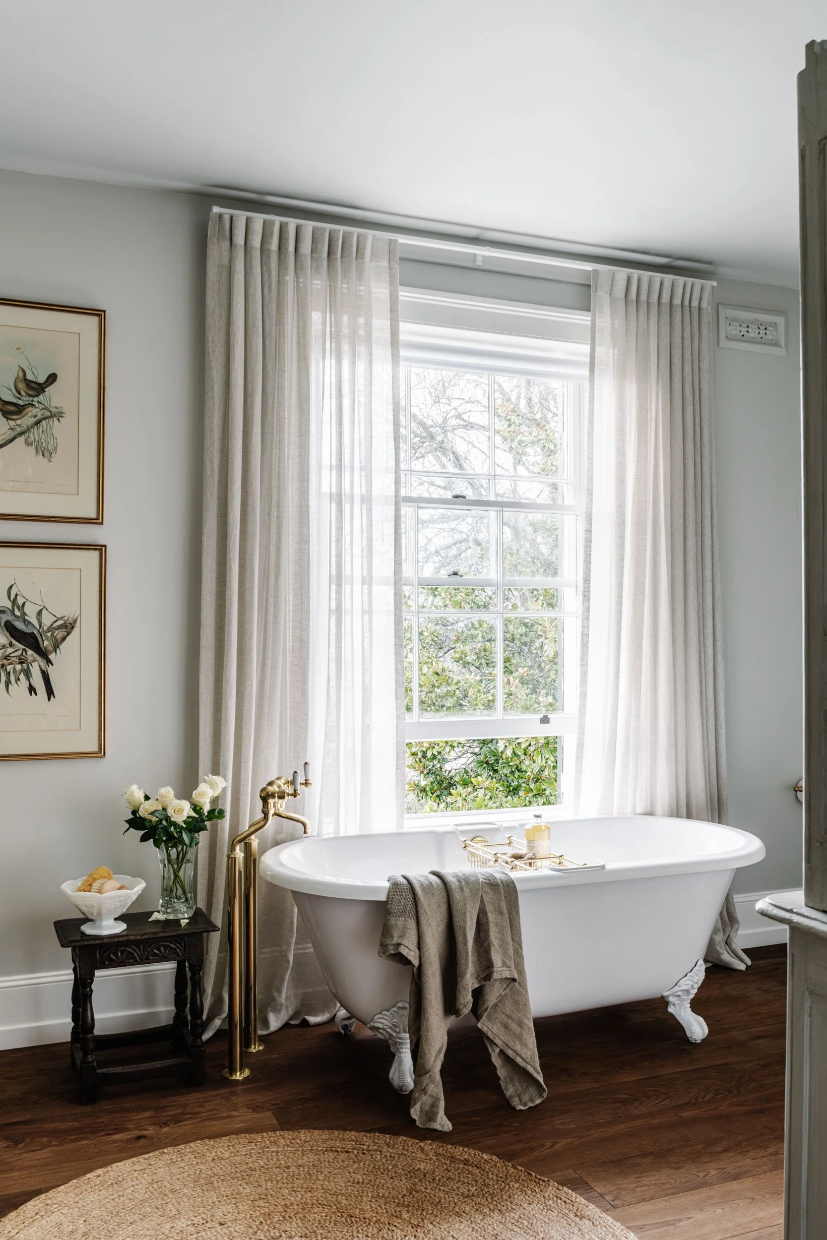 A bright bathroom with a white clawfoot bathtub placed in front of a large window, with cream curtains partly drawn, a small dark wooden side table with a white bowl of flowers, and wall art featuring bird illustrations.