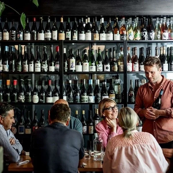 People seated around a table in a wine bar with shelves of wine bottles behind them, and a man holding a wine bottle standing nearby.