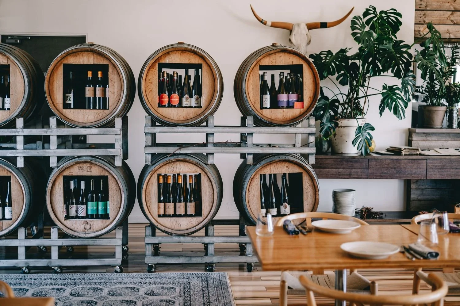 Room with wine bottles stored inside wine barrels, a mounted animal skull with horns on the wall, large potted plants, and a wooden dining table set with plates and utensils.