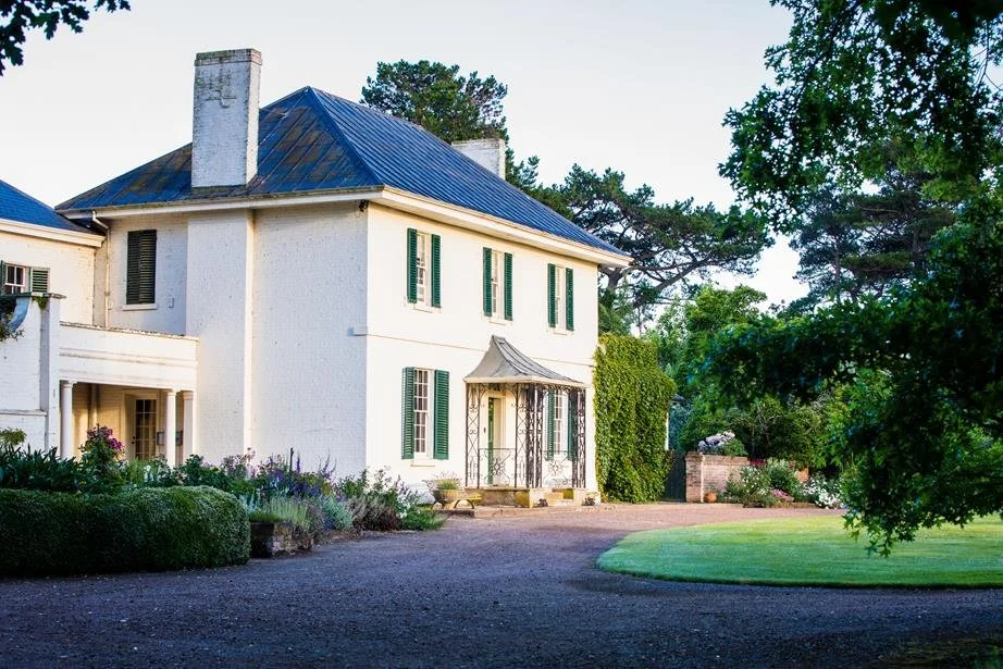 Large white two-story house with green shutters, a blue metal roof, and a small front porch with decorative ironwork, surrounded by trees and a well-maintained lawn.