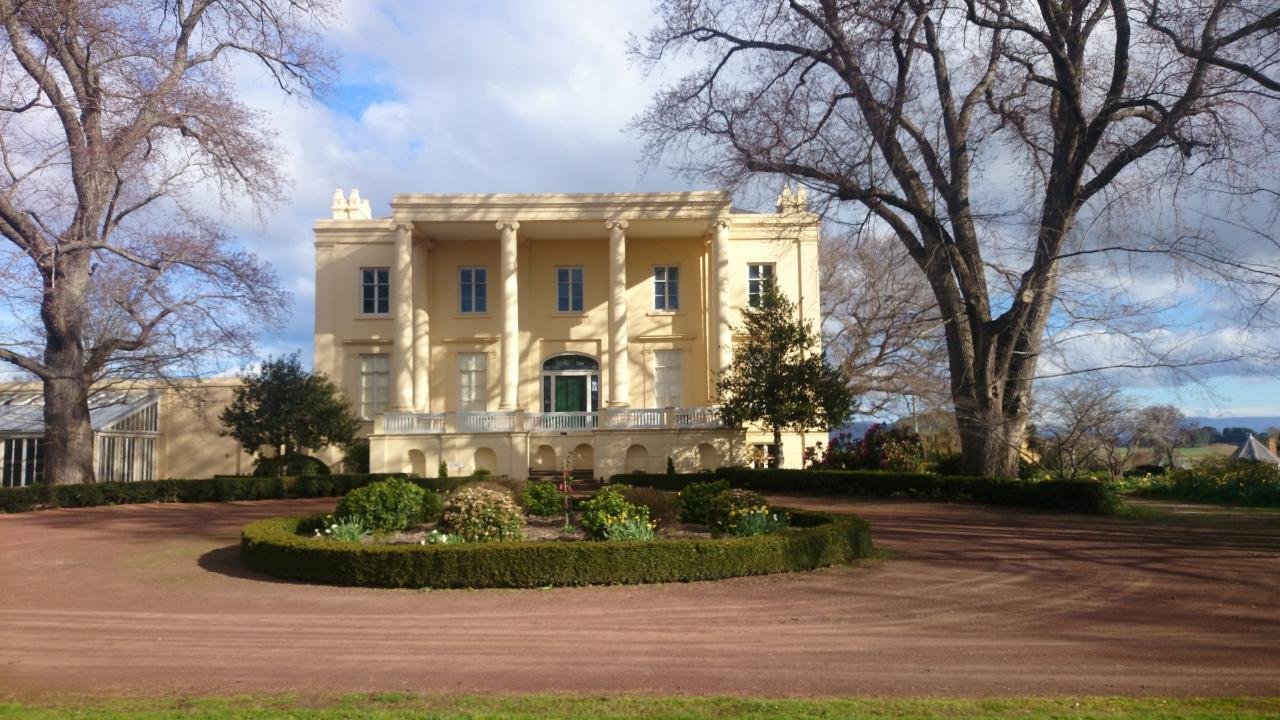 A large, elegant, cream-colored mansion with four columns at the front entrance, surrounded by trees and landscaped gardens, under a partly cloudy sky.