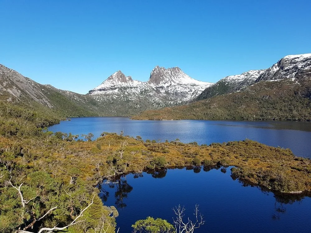 Scenic view of a mountain lake with snow-capped peaks, surrounded by green forest and clear blue sky.