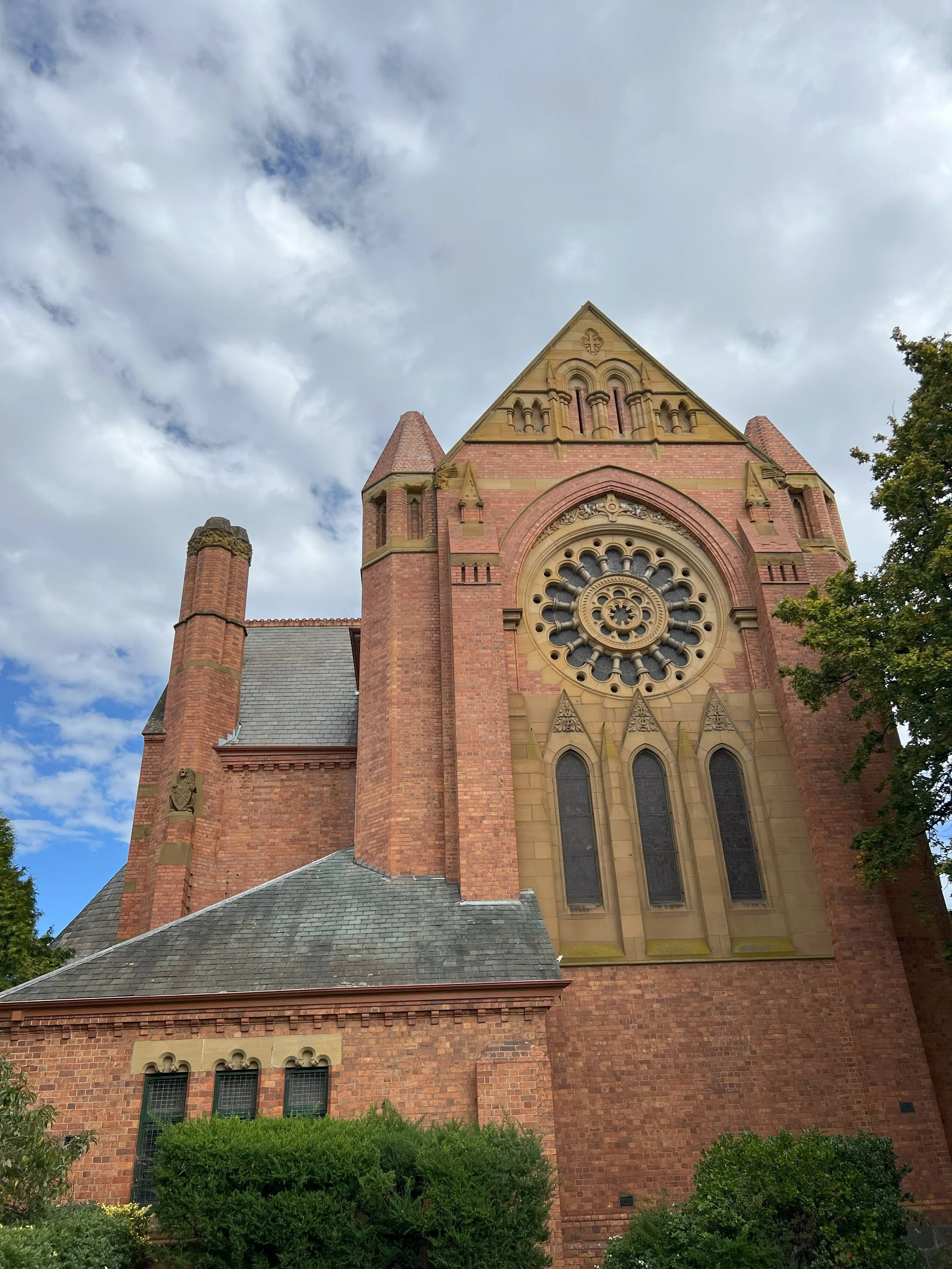 A tall, historic red brick church with a large circular stained glass window and pointed arches, set against a partly cloudy sky with some trees in the foreground.