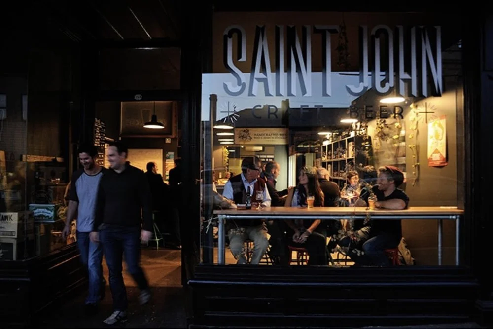 People sitting at tables and walking inside Saint John Craft Beer restaurant, with the interior and bar area visible through the window.