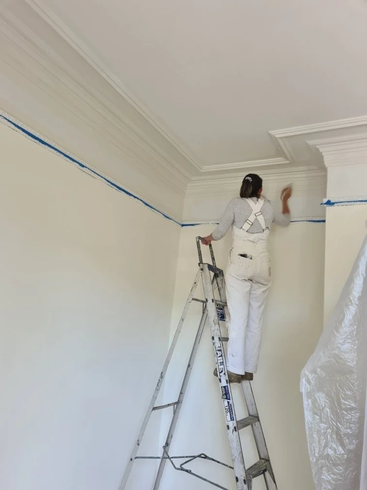 A woman painting the ceiling at Beulah House while standing on a step ladder, dressed in white overalls and a grey long-sleeve shirt, carefully restoring the Georgian room to its original beauty.