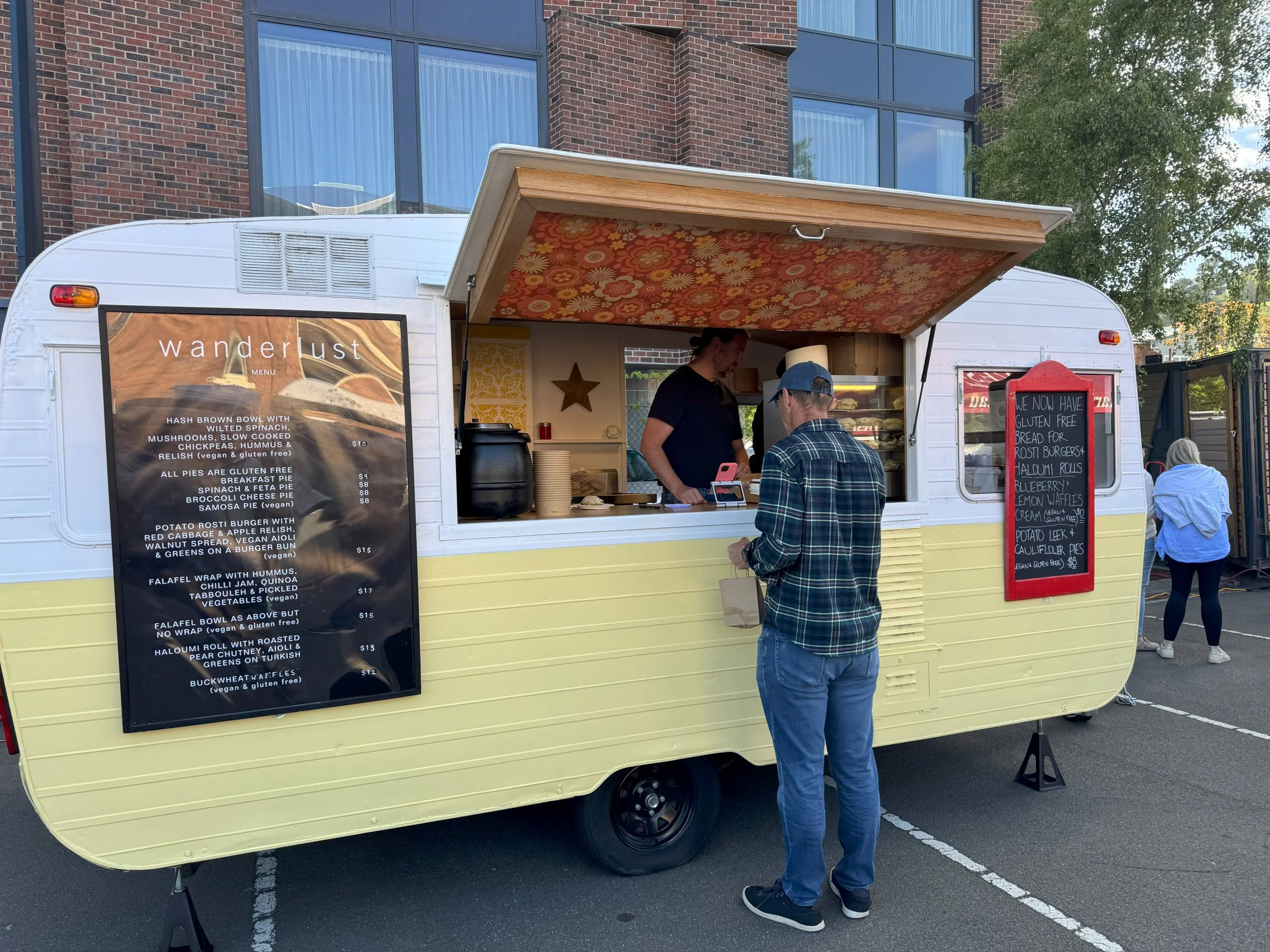 A food truck named Wanderlust parked in a lot with a brick apartment building in the background. The truck has a black menu board and a chalkboard with handwritten menu items. A customer stands at the window ordering food, and a woman inside is preparing the order.