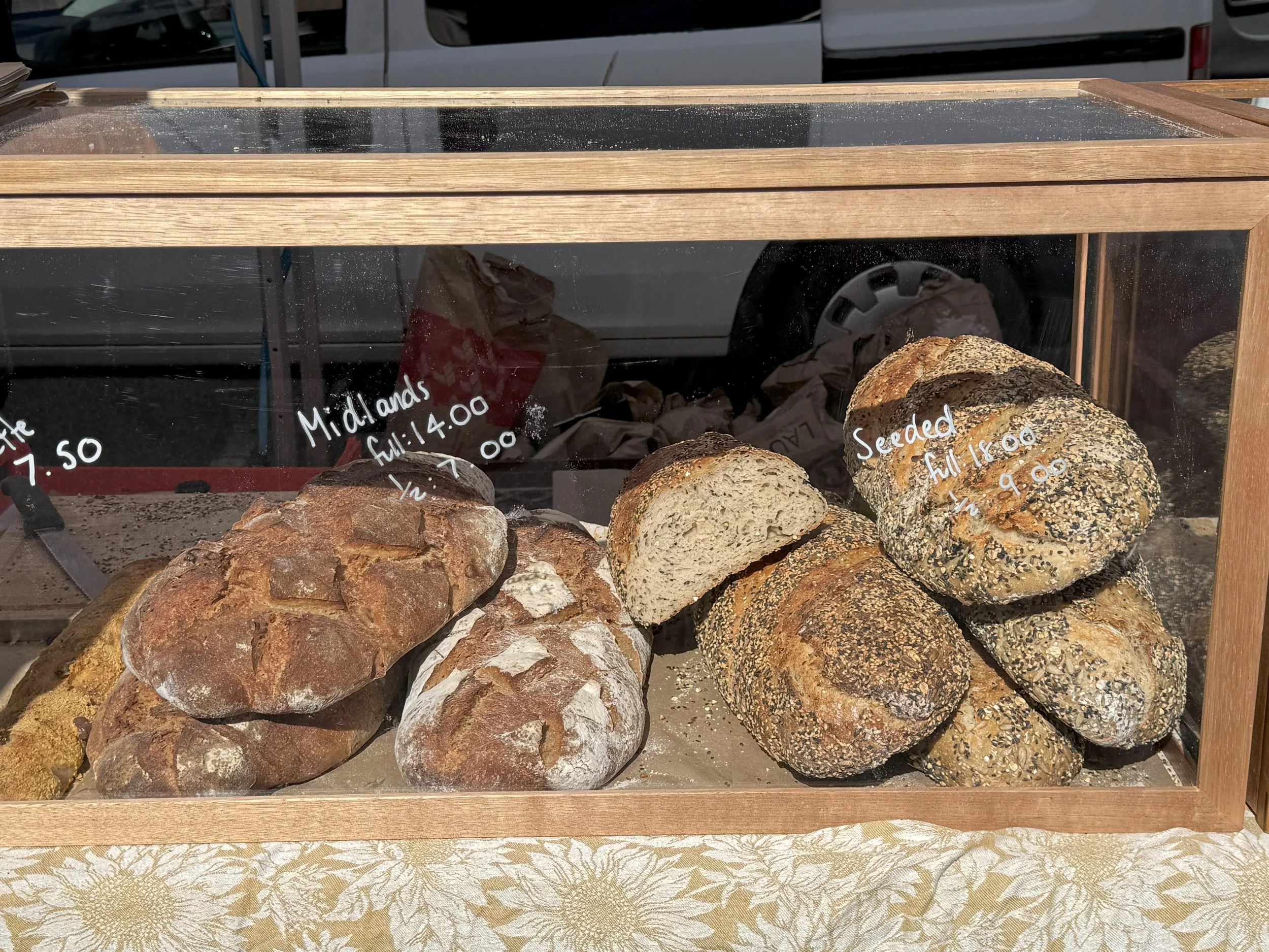 Assorted bread loaves displayed in a glass bakery case, labeled with prices and types including seeded and various artisan varieties.