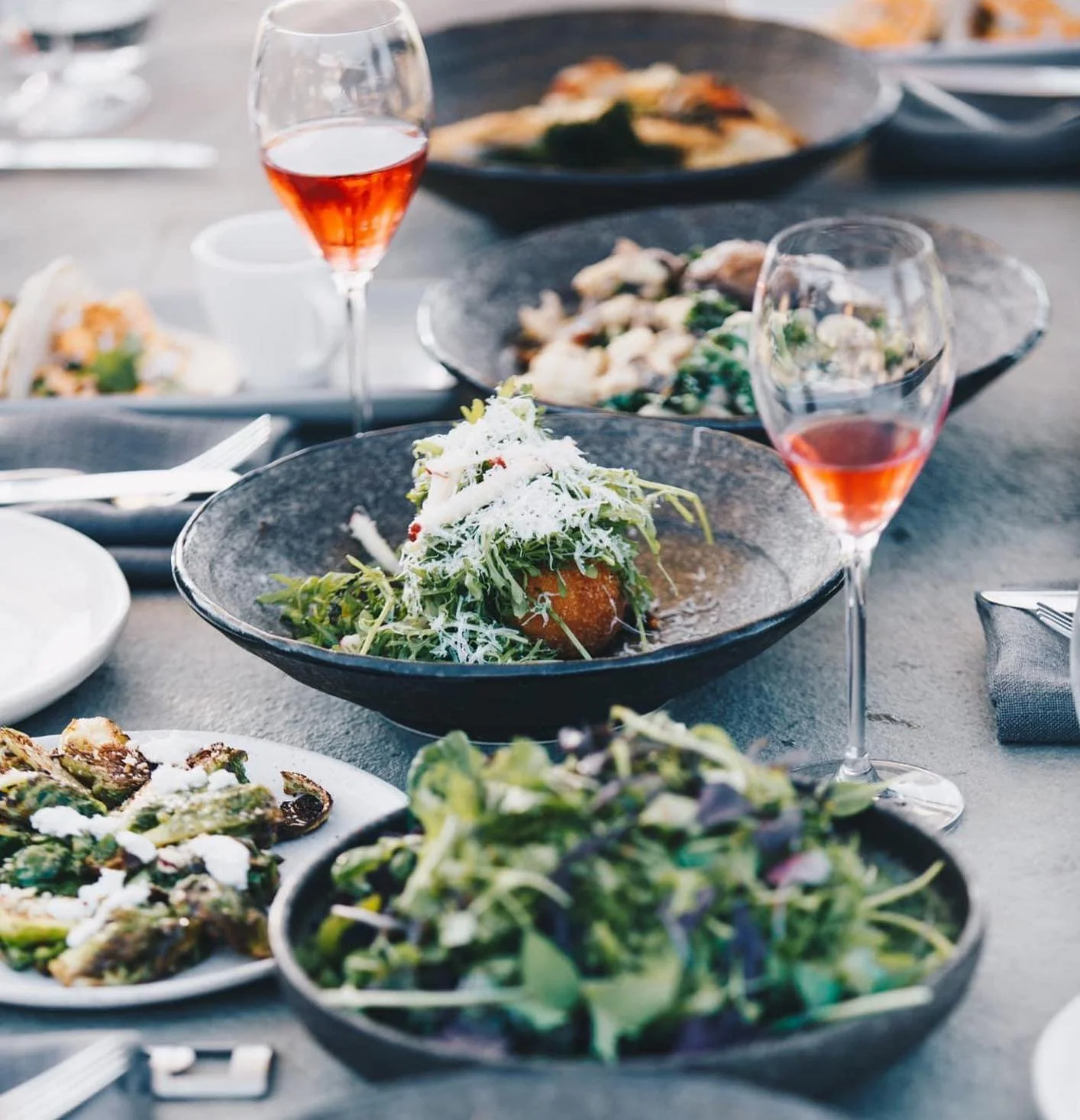 A table set with various dishes including salads, a plate of grilled vegetables, multiple bowls of salads, and two glasses of rosé wine.