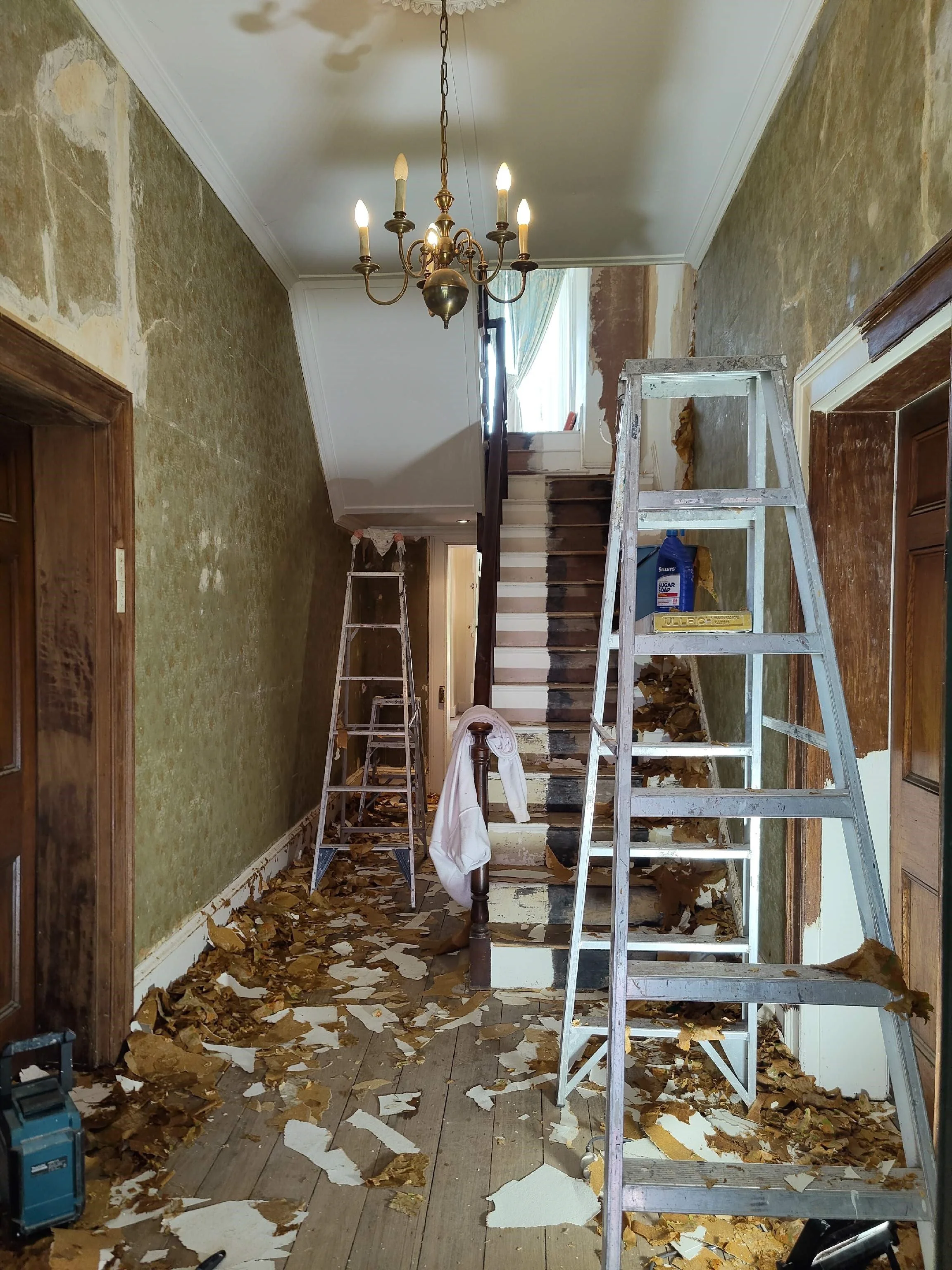 An interior restoration scene at Beulah House showing damaged walls, stairs, and ceiling with debris on the floor, two ladders, a chandelier, and tools scattered during the meticulous Georgian-era renovation.