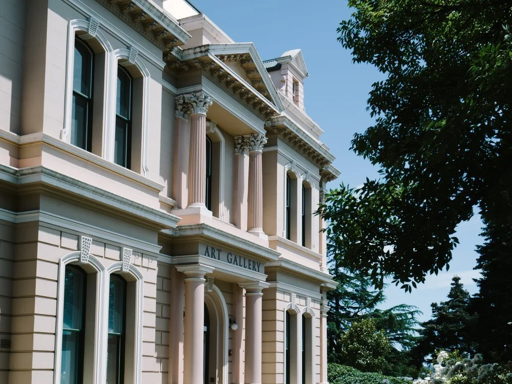 A historic art gallery building with a classical architectural style, featuring tall windows, columns, and decorative details, surrounded by trees.