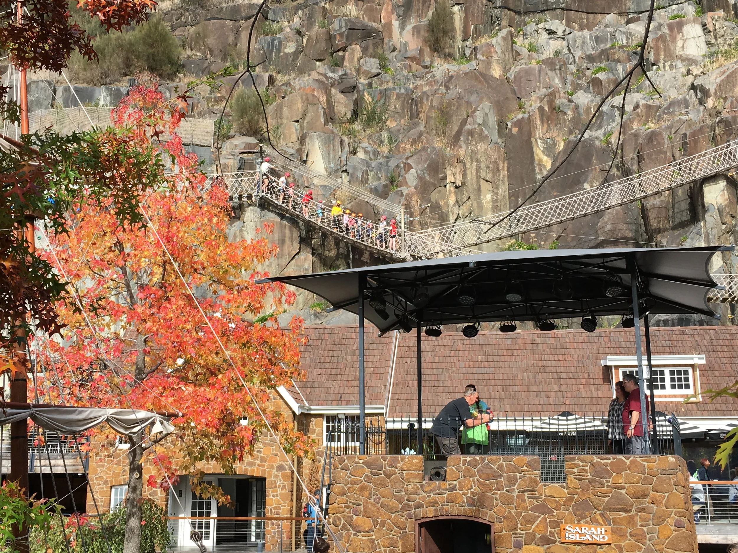 A stage with people gathering under a canopy, a colorful tree with red autumn leaves, stone buildings, and a mountainous rocky background with a bridge and tourists walking on it.