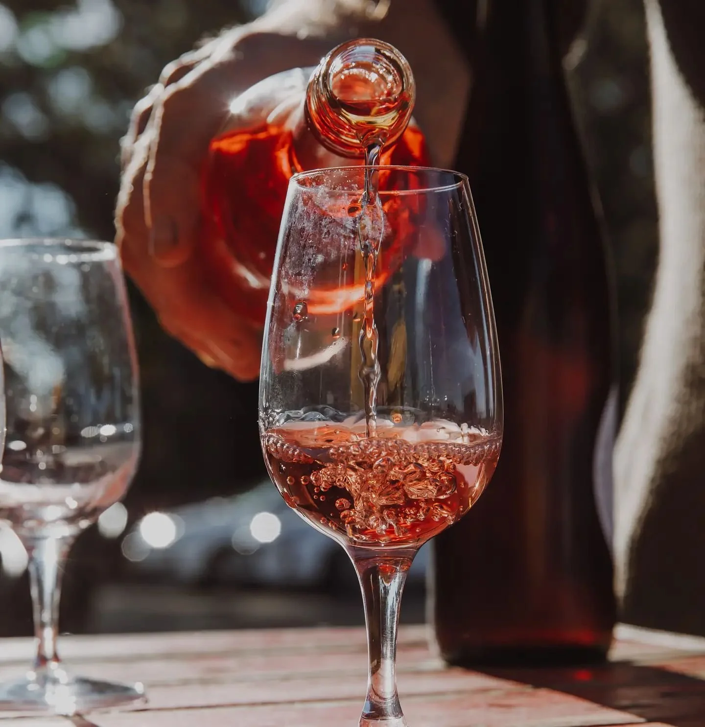 A person in a dark jacket pours rosé wine into a wine glass outdoors with blurred background and sunlight.