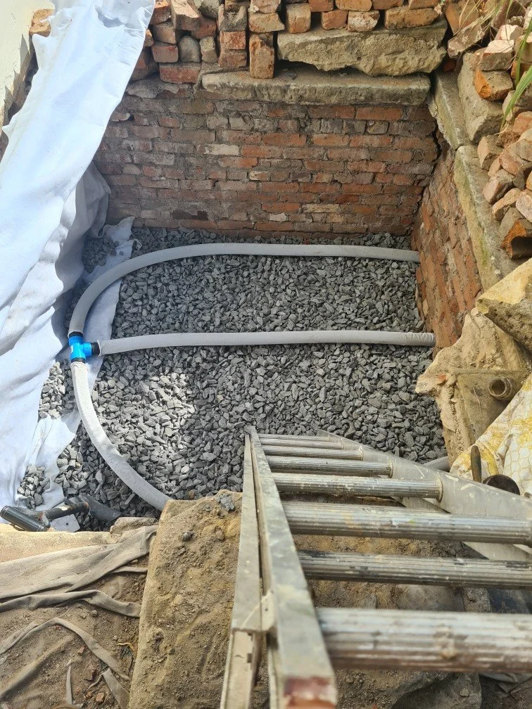 Construction site with a brick wall, gray gravel floor, and plastic tubing for plumbing or wiring. A metal ladder leans against the edge of the excavation.