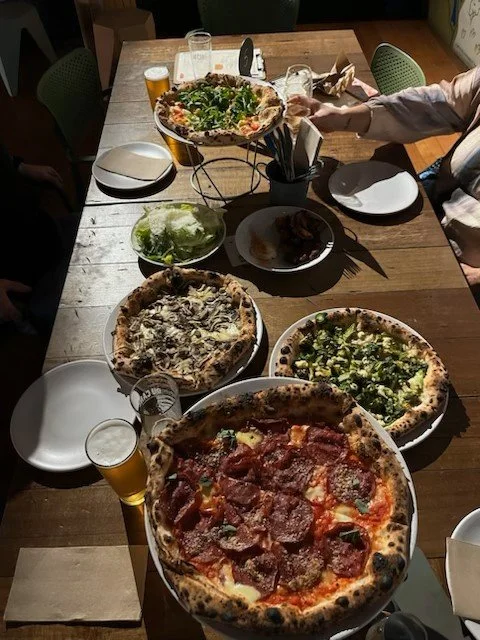 Pizza on the table with various toppings, salad, glasses of beer, napkins, utensils, and a person reaching with a plate in a restaurant setting.