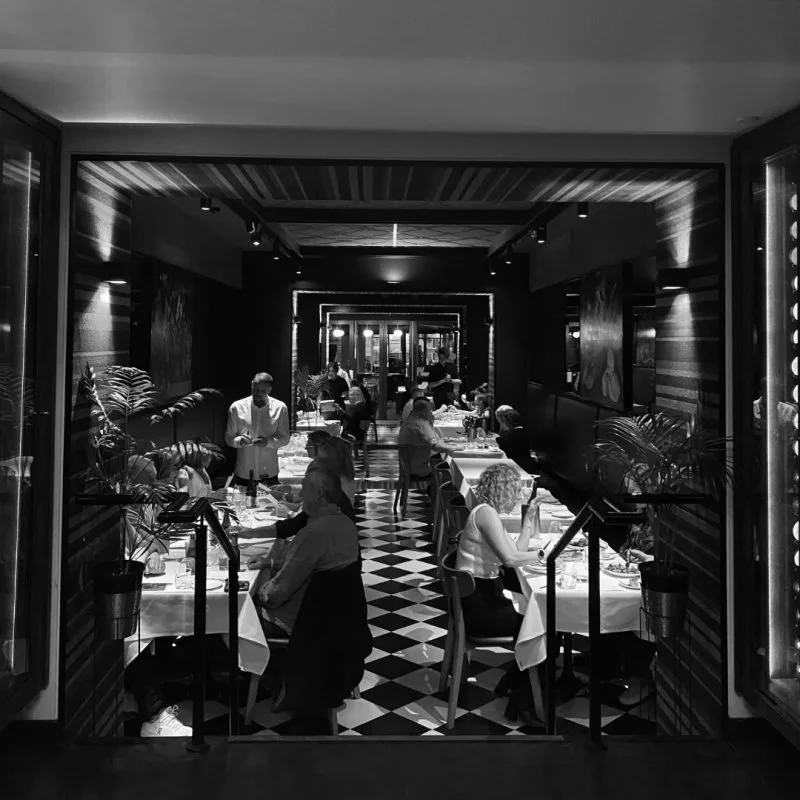Black and white photo of a restaurant interior with diners seated at tables, waitstaff serving, and a checkered floor.