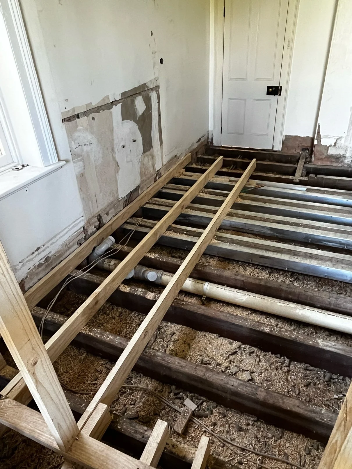 Room with subflooring being replaced, showing exposed floor joists and pipes, unfinished wall, and a door.