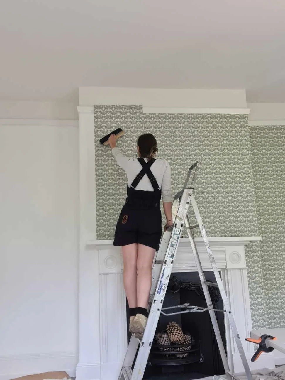 A woman carefully cleaning the patterned wallpaper above a white fireplace mantel at Beulah House, standing on a ladder and restoring the Georgian interior with delicate attention to detail.