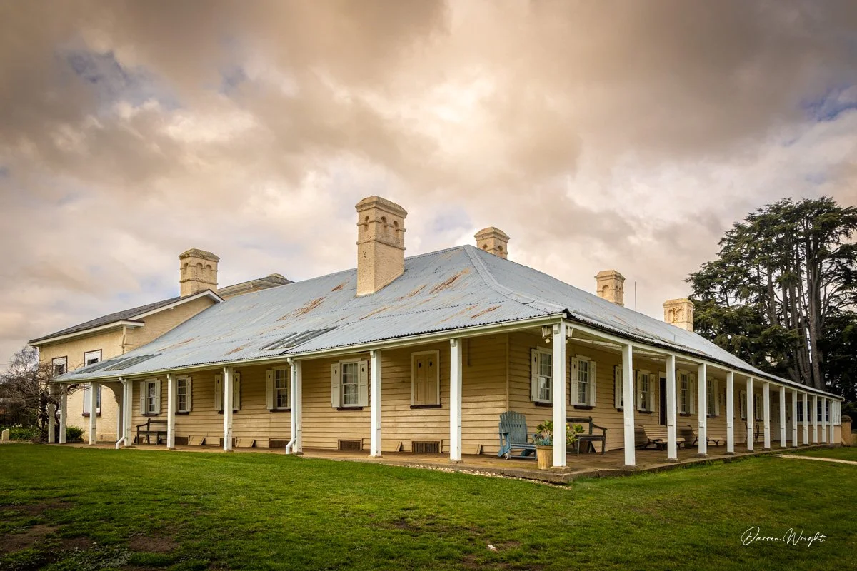 A historic wooden house with a corrugated metal roof, multiple chimneys, and a covered porch with benches and potted plants, set against a cloudy sky and surrounded by green grass and trees.