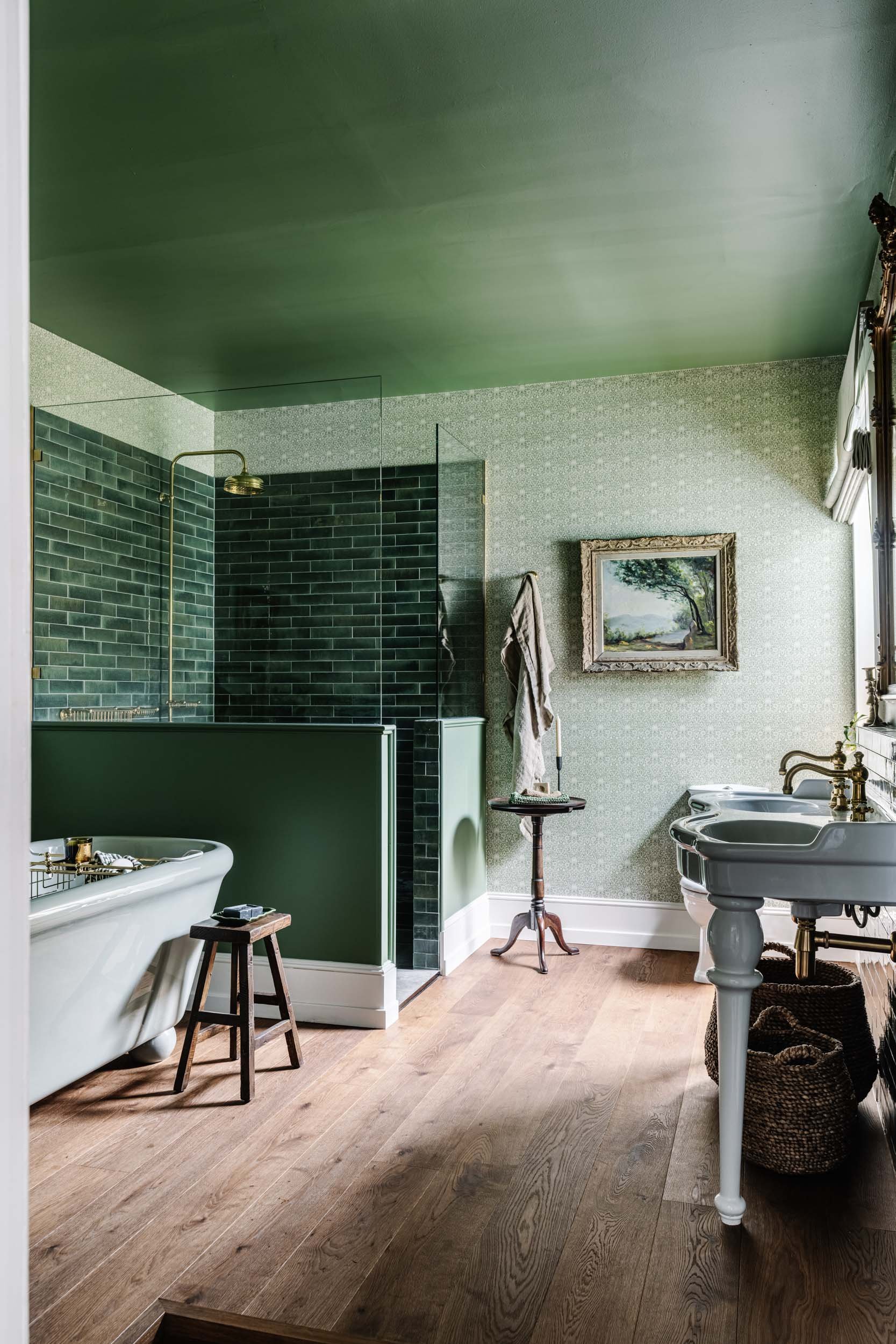 A vintage bathroom with a clawfoot bathtub, green tiled shower, and double sinks with brass fixtures. The room features wood flooring, patterned wallpaper, a framed landscape painting, and a window with ornate drapes.