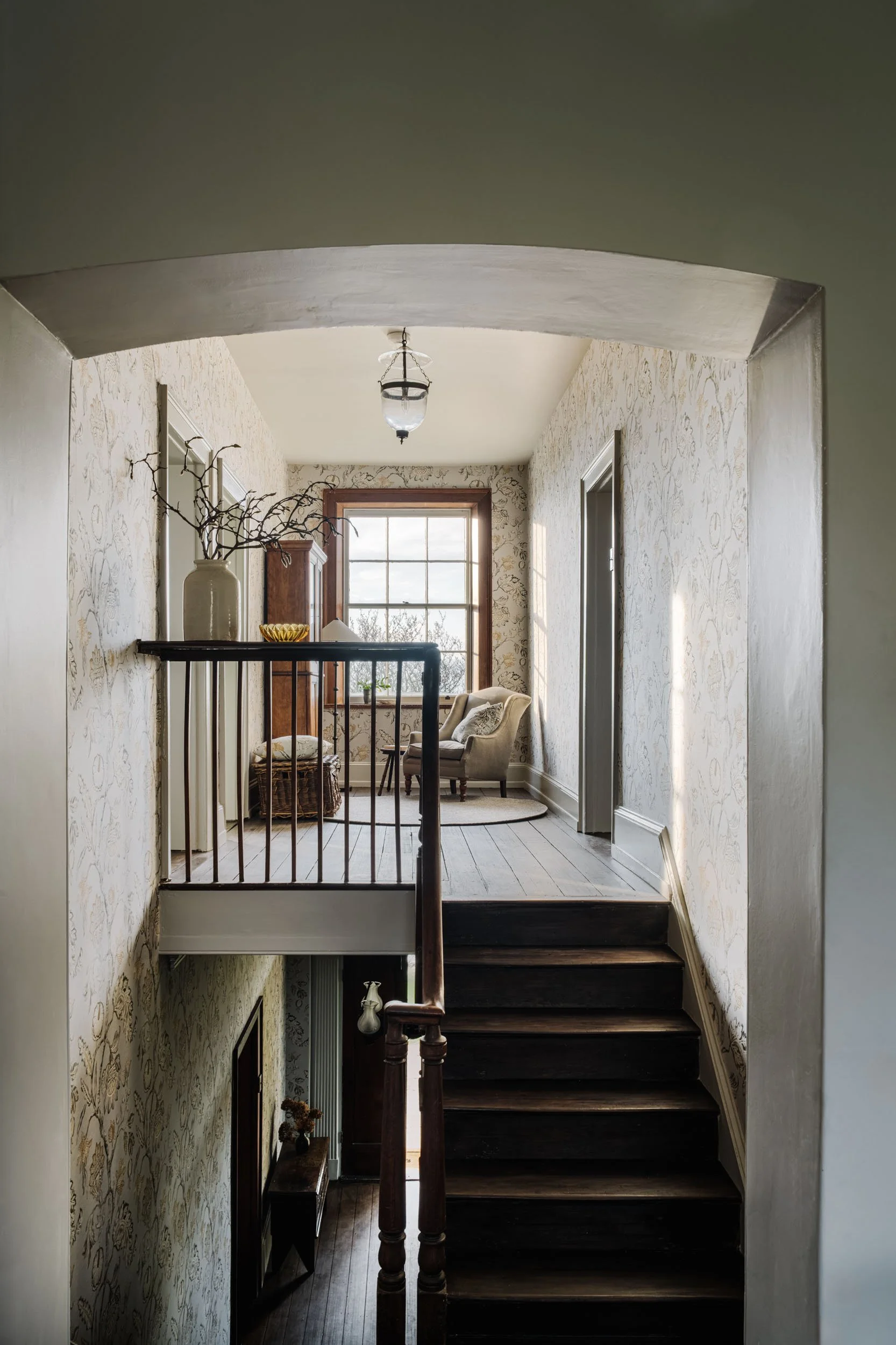 View of a vintage-style stairway leading to a small sitting area with a large window, patterned wallpaper, a beige armchair, and decorative items.