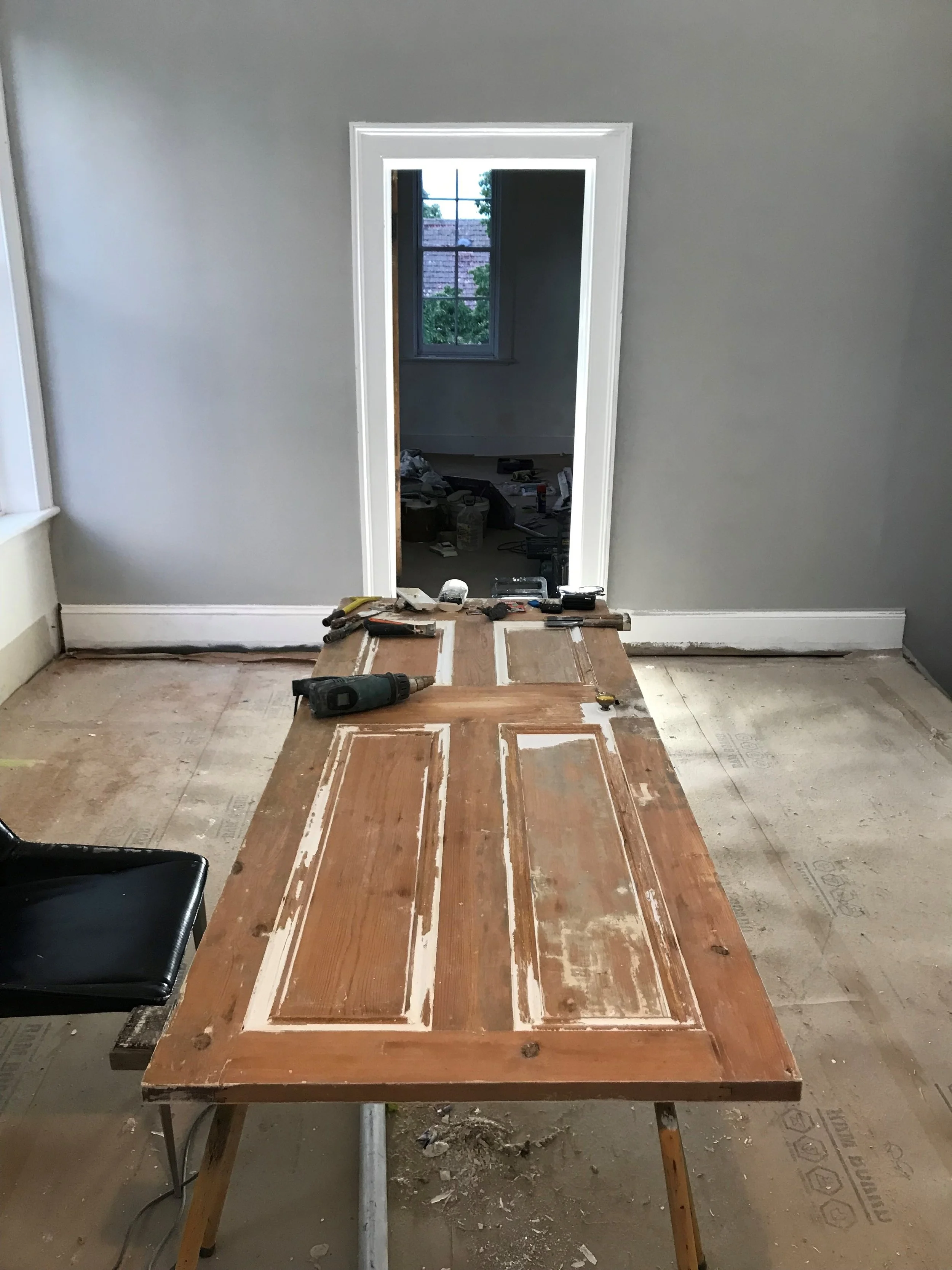 Interior room under renovation with a wooden door lying on a worktable, various tools on the table, and an entryway leading to another room with a window.