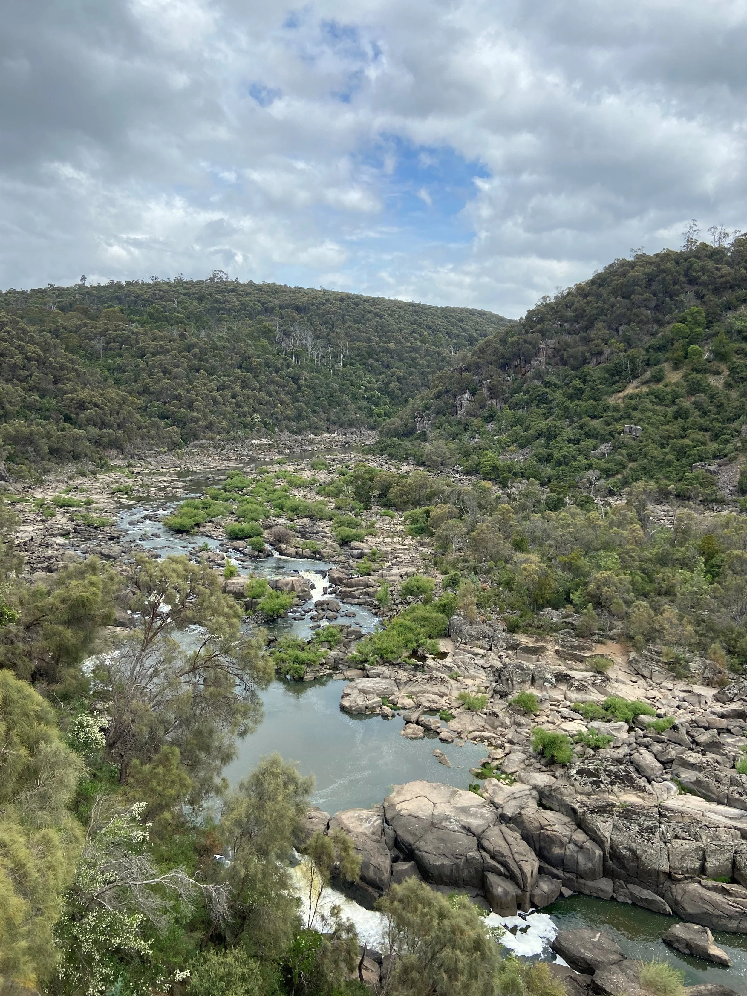 A scenic view of a river flowing through a rocky valley surrounded by green hills under a partly cloudy sky.