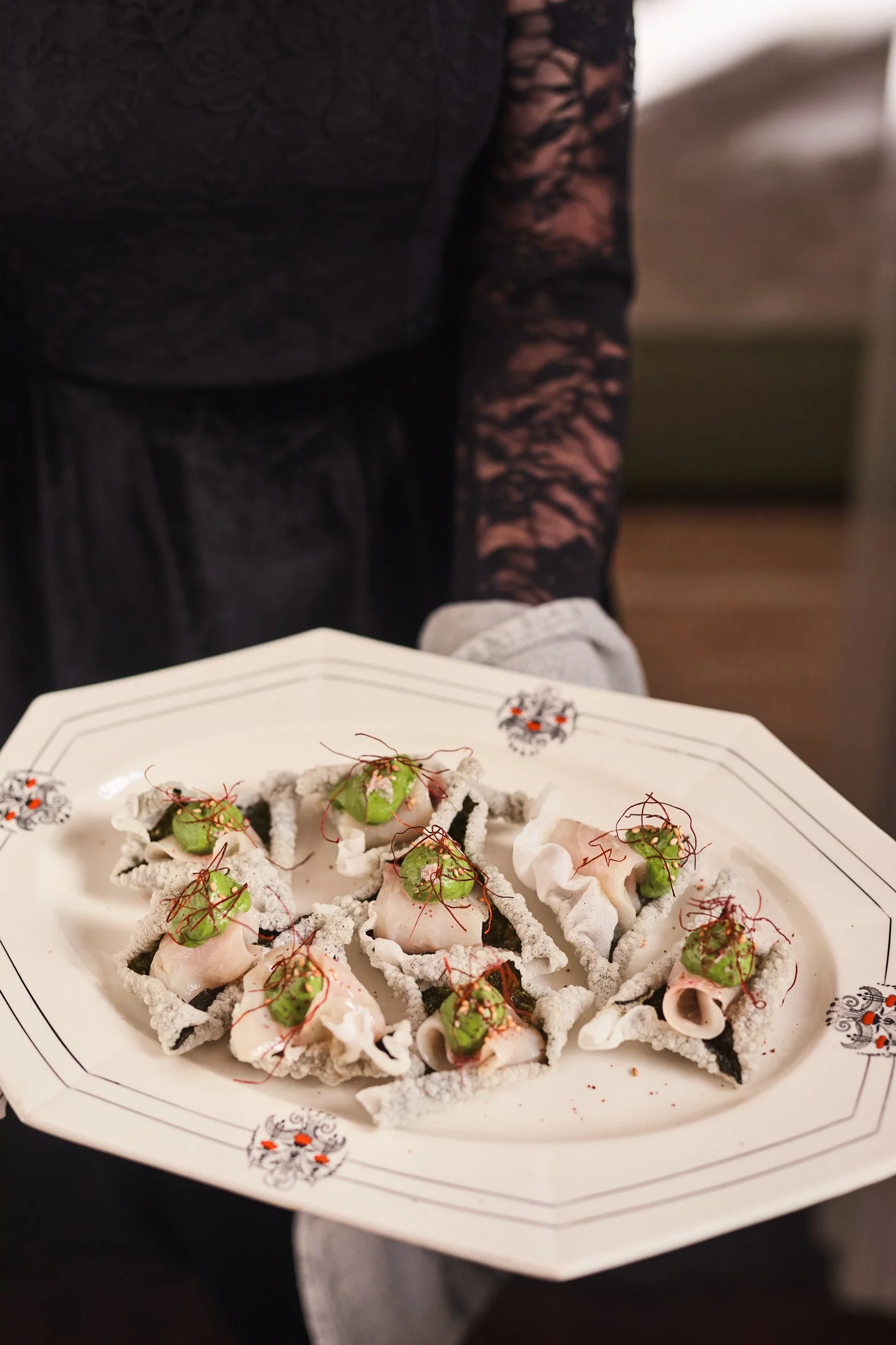 A guest at Beulah House wearing black lace holds a decorative white plate of small appetizers topped with green sauce and red microgreens, reflecting the home’s refined Georgian hospitality.