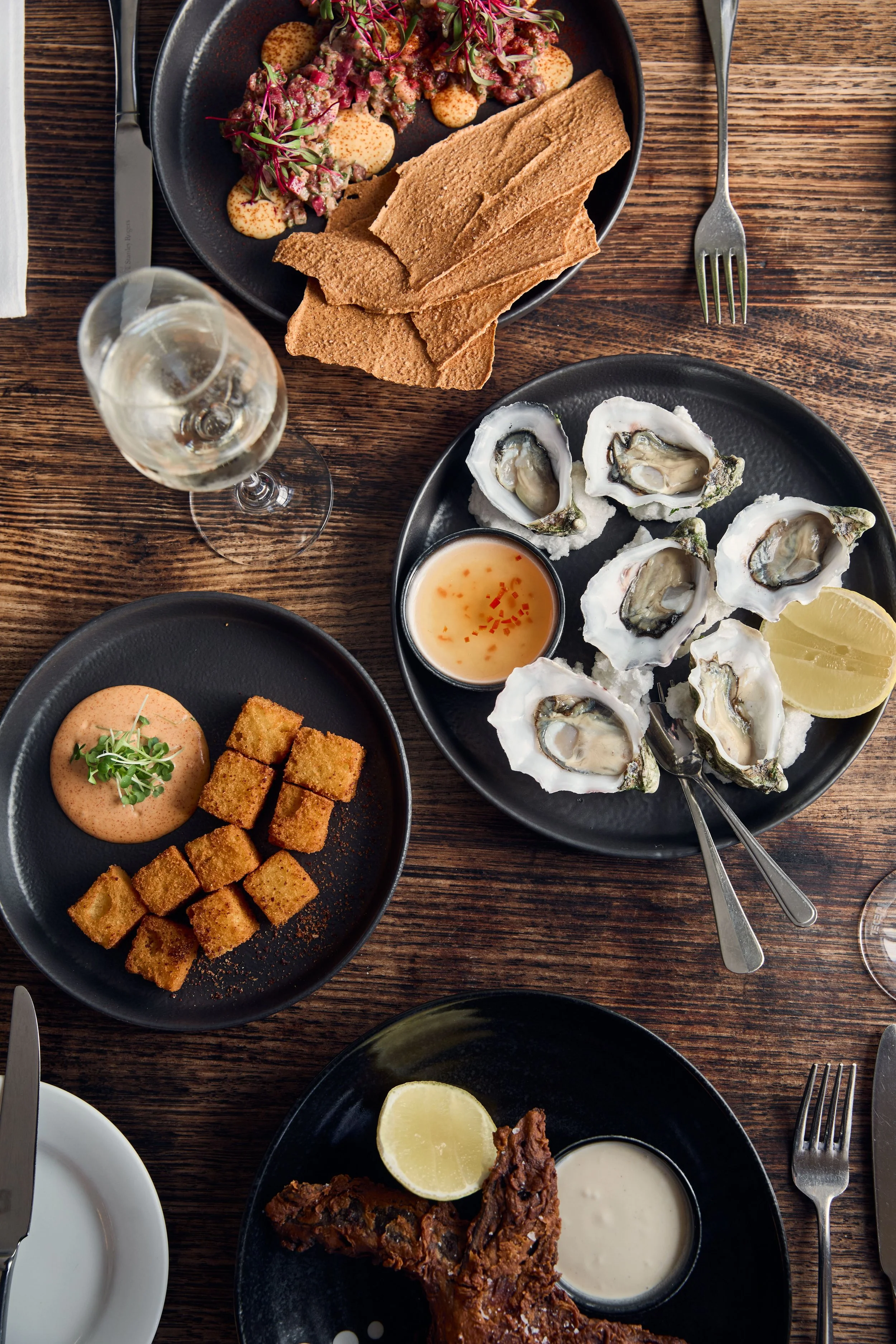 A dining table with various dishes including oysters on a black plate with lemon wedges and dipping sauce, a black plate with fried chicken and dipping sauce, a black bowl with cubed fried food and pink sauce, a black bowl with a salad and bread slices, and a glass of white wine.