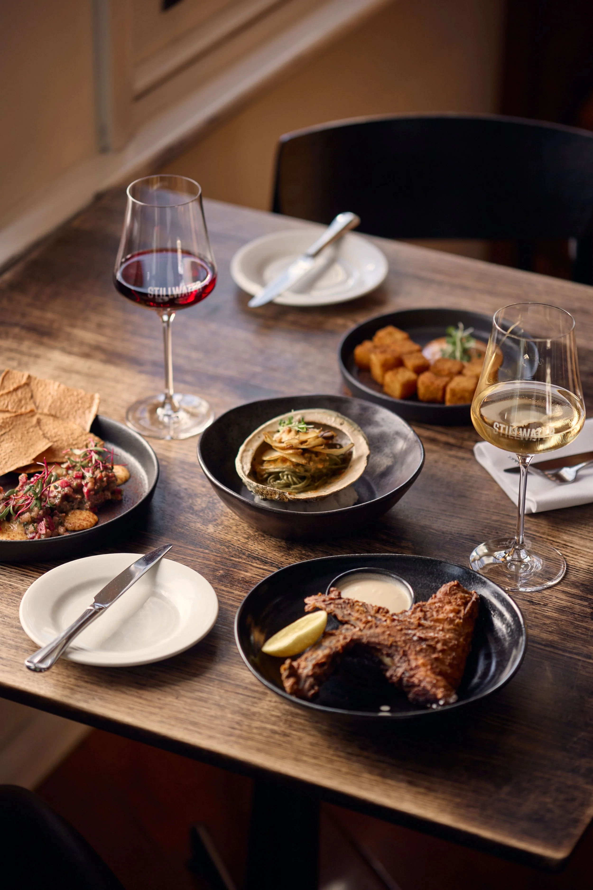 A wooden table set with various dishes including a cooked steak with lemon, a shellfish, and small fried items, along with two glasses of white and red wine, and some plates and cutlery.