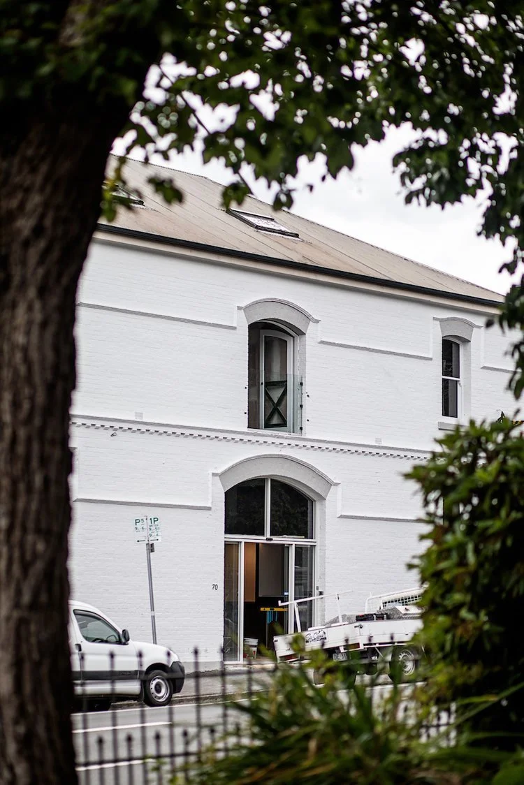 A white brick building viewed through trees and a fence, with a vehicle and a small boat parked outside.