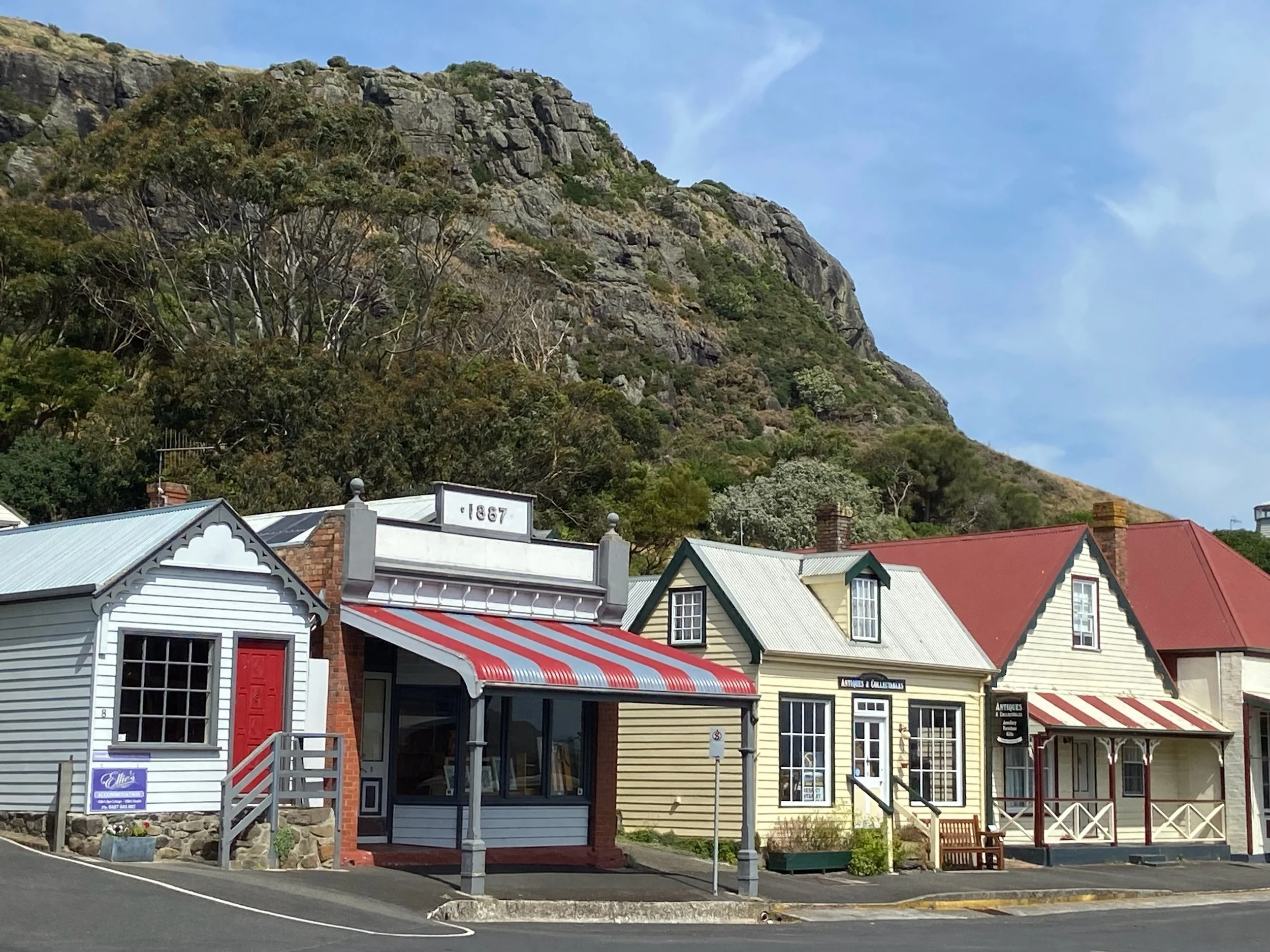 Colorful shops with a mountain and trees in the background, clear sky, and a sidewalk in front.