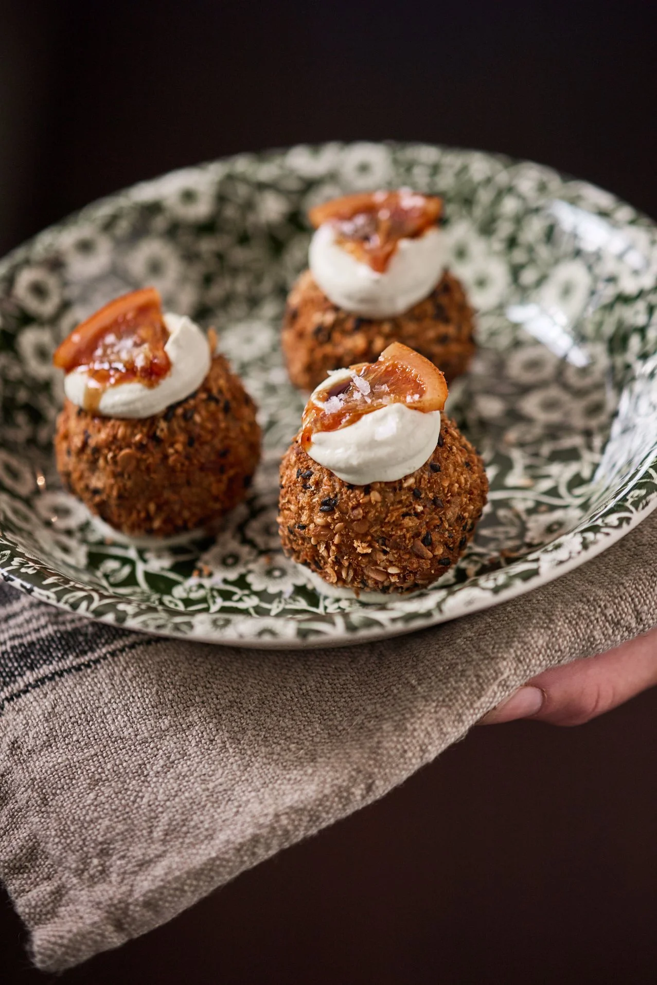 Plate of three baked dessert balls topped with whipped cream, caramel sauce, and a small piece of caramelized fruit, on a dark background.