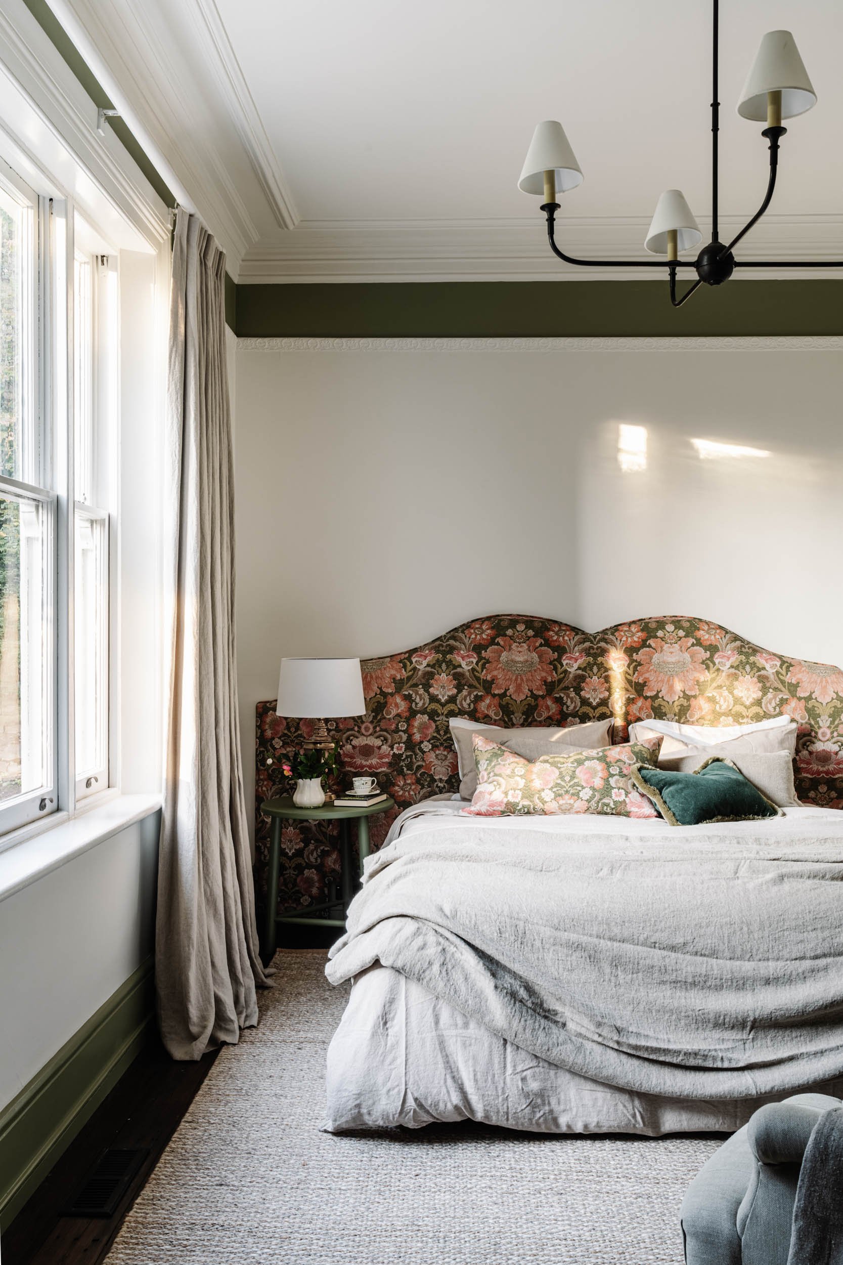 A bedroom with a window in the left, beige curtains, a bed with an ornate floral headboard, white bedding, and decorative pillows. A small bedside table with a lamp, flowers, and a cup is next to the bed. Light streams through the window, illuminating part of the room.
