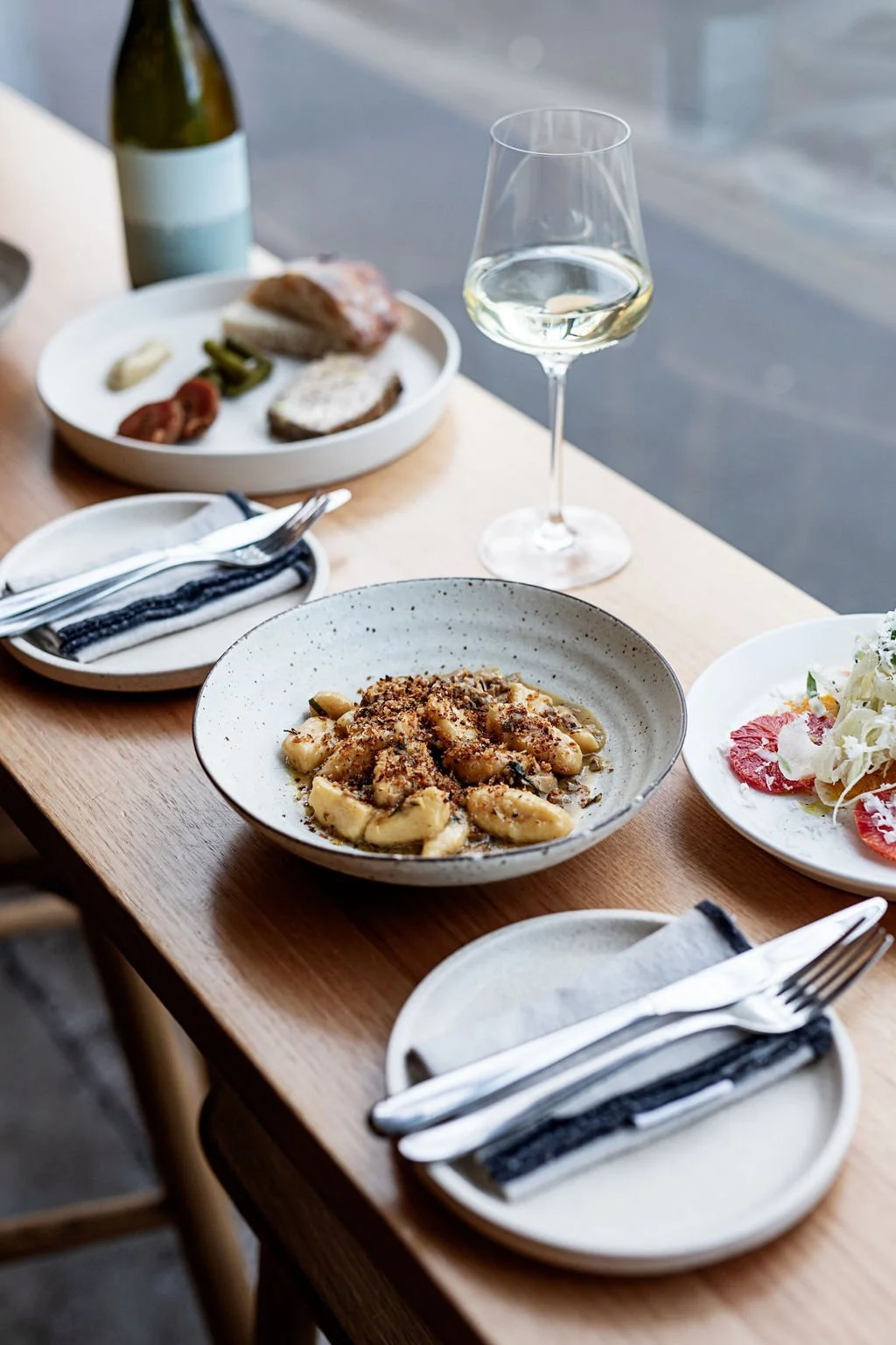A dining table set with assorted foods including a glass of white wine, a plate of pasta with cheese, a salad, and a charcuterie board with meats and cheeses. Plates, utensils, and napkins are also on the table.
