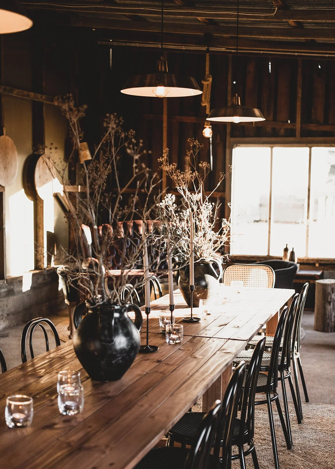 A rustic dining room with a long wooden table, black chairs, and large black vases filled with dried branches as centerpieces. Warm lighting from pendant lamps and sunlight streaming through large windows.